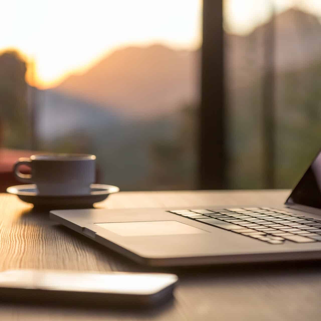 A laptop with a cup of coffee on a wooden table at sunset
